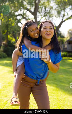 Madre che dà alla figlia un giro in piggyback, sorridendo e divertendosi all'aperto con la famiglia Foto Stock