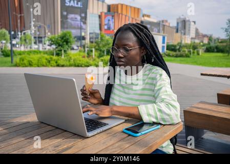 La donna afroamericana in occhiali ama il cono gelato mentre lavora su un computer portatile al tavolo all'aperto in un parco urbano, combinando svago e produttività. Freelance nomadi digitali in natura. Foto Stock