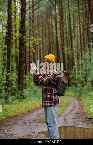 L'escursionista riccia con berretto giallo e camicia in flanella scatta foto con lo smartphone mentre cammina attraverso la lussureggiante foresta verde Foto Stock