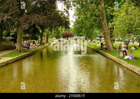 Persone che si rilassano lungo il pittoresco fiume Windrush nel villaggio Cotswold di Bourton on the Water, Gloucestershire, Inghilterra, Regno Unito Foto Stock