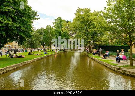 Persone che si rilassano lungo il pittoresco fiume Windrush nel villaggio Cotswold di Bourton on the Water, Gloucestershire, Inghilterra, Regno Unito Foto Stock