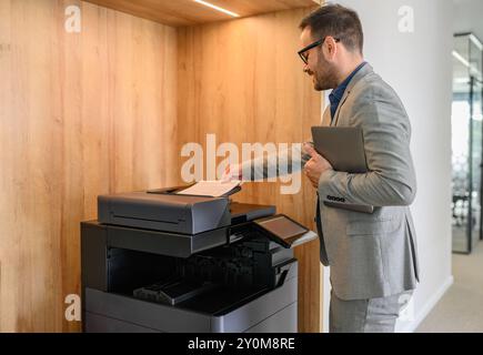 Vista laterale di un professionista che tiene un computer portatile e inserisce la carta nella fotocopiatrice in un ufficio moderno Foto Stock