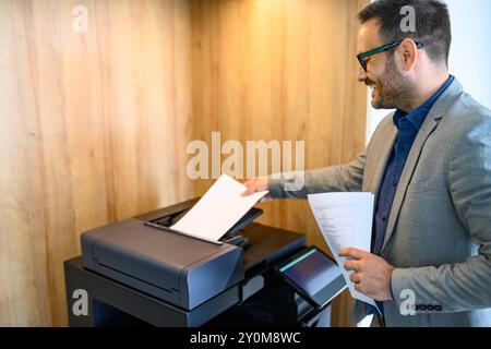 Vista laterale di un giovane uomo d'affari sorridente che mette le carte nella fotocopiatrice in un ufficio moderno Foto Stock