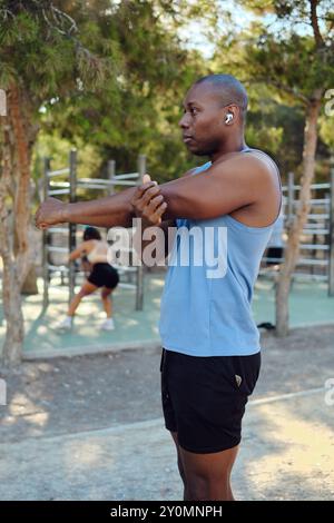 Atletico africano vestito con camicia blu senza maniche, pantaloncini neri fanno stretching delle spalle e scaldano gli esercizi nel parco, nella soleggiata giornata estiva. Sport, guarisci Foto Stock