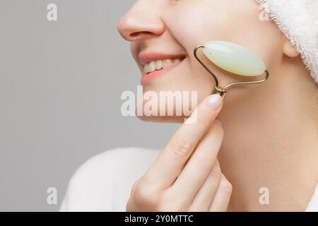 Una donna con l'abito bianco e un asciugamano sulla testa sorride tenendo i rulli facciali in giada sotto gli occhi. concetto di levigatura delle rughe Foto Stock