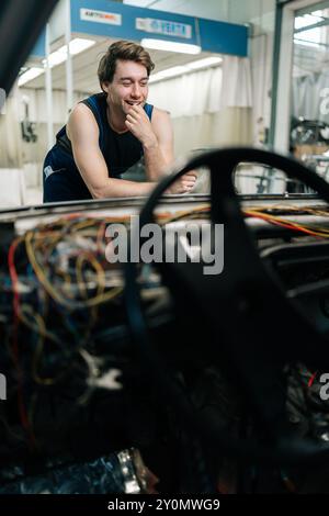 Scatto verticale di un allegro meccanico sorridente mentre si pensa a come riparare l'impianto elettrico di un'auto appoggiandosi al cofano dell'auto. Ridendo perplesso Foto Stock