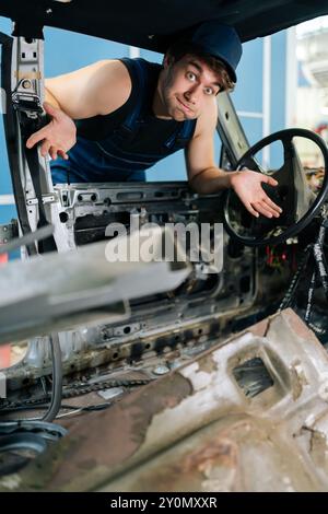 Ritratto verticale del movimento maschile meccanico sconcertato all'interno di un'auto smontata, che mostra l'esperienza nel settore automobilistico e i lavori in corso, guardando la fotocamera Foto Stock