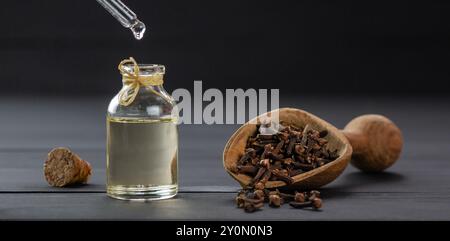 Bottiglia in vetro di olio essenziale di chiodi di garofano e chiodi di garofano in pala di legno o cucchiaio su un tavolo di legno nero, gocciolamento di olio Foto Stock