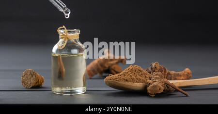 Bottiglia in vetro di olio essenziale galangal, radice e polvere di galanga in cucchiaio di legno su tavolo di legno nero, (Alpinia officinarum ), gocciolamento di olio Foto Stock