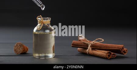 Bottiglia di olio di cannella in vetro con bastoncini di cannella su fondo di legno nero, gocciolamento di olio Foto Stock