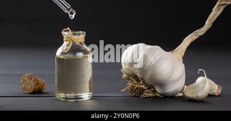 Aglio maturo e crudo e olio all'aglio in un bicchiere di bottiglia su un tavolo di legno nero, gocciolamento di olio Foto Stock
