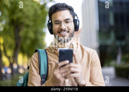 Giovane con le cuffie che usa lo smartphone all'aperto. Goditi la musica indossando un abbigliamento casual con lo zaino. Mostrare relax, godersi la giornata. Cattura lo stile di vita, la tecnologia moderna e la vita degli studenti. Foto Stock