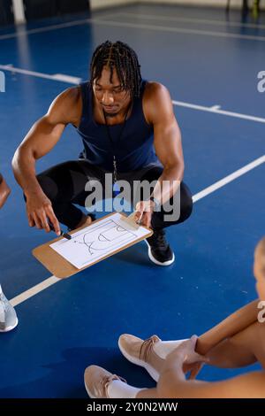 Allenatore maschile che spiega la strategia di basket sugli appunti alle giocatrici in palestra Foto Stock