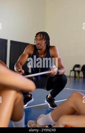 Allenatore maschile che tiene gli appunti e dà istruzioni alle giocatrici di basket in palestra Foto Stock