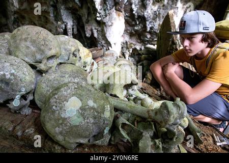 Il turista dei bambini esplora il sito di sepoltura Lombok Parinding a Tana Toraja, teschi umani, ossa e bare di legno nella spettacolare tomba della grotta, Sulawesi Foto Stock