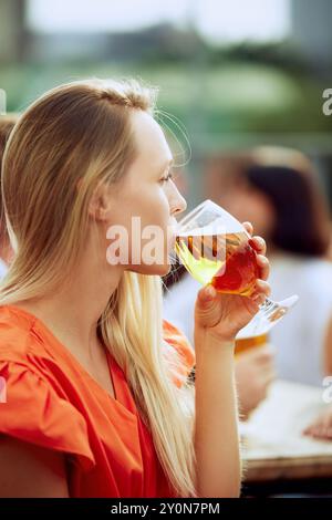 Foto ravvicinata di una giovane donna bionda seduta in terrazza e che beve birra artigianale fredda il venerdì sera sulla terrazza del caffè. Foto Stock