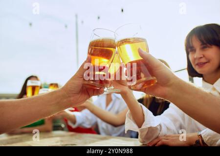 Gli amici si riuniscono intorno al tavolo, alzando i bicchieri con un brindisi per passare i momenti migliori. L'atmosfera vivace e le risate condivise rendono perfetta la giornata di sole. Foto Stock