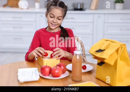 Una ragazza carina che mette i pomodori nel cestino da pranzo al tavolo di legno in cucina. Prepararsi per la scuola Foto Stock