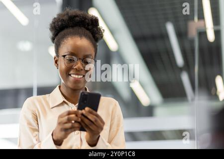 Donna d'affari afro-americana sorridente mentre utilizza lo smartphone in un moderno ambiente d'ufficio. L'immagine cattura l'interazione professionale con la tecnologia digitale, sottolineando l'umore allegro Foto Stock