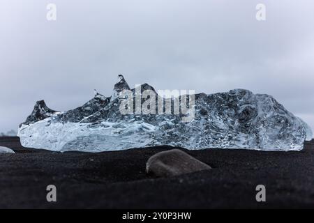 Iceberg trasparente adagiato sulla sabbia nera vulcanica di Diamond Beach in Islanda, vista ravvicinata dall'angolo basso con cielo nuvoloso sullo sfondo. Foto Stock
