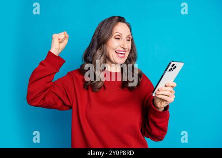 Foto di una signora piuttosto anziana che usa il telefono alzare il pugno indossare un pullover rosso isolato su sfondo di colore blu Foto Stock