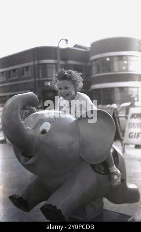 1960 anni, una giovane ragazza eccitata fuori su una passeggiata sul lungomare seduta su un giro di divertimento "elefante". Foto Stock