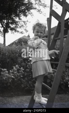 1950, storica, una bambina che posa per una foto sul gradino di uno scivolo in legno nel suo bel vestito da scuola domenicale, giacca, gonna a pieghe e arco di capelli. Foto Stock