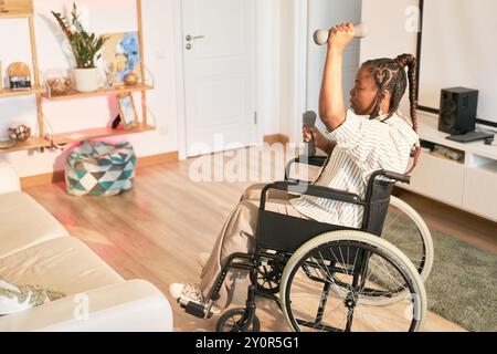 Persona su sedia a rotelle che solleva manubri in un ambiente domestico pieno di vari oggetti per la casa. L'arredamento include piante e decorazioni circonda l'area senza utilizzare attrezzature professionali Foto Stock