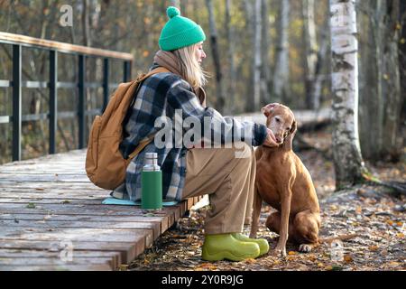 La donna che viaggia fa una pausa per l'escursione, dà da mangiare a mano il cane da caccia Vizsla, seduto su un sentiero ecologico nel parco autunnale Foto Stock