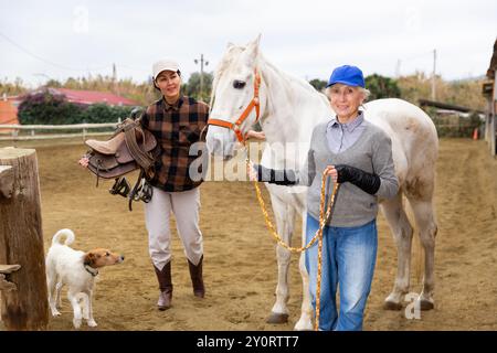 Anziana stalla donna che conduce cavallo a cavallo all'arena di equitazione all'aperto, mentre donna asiatica che tiene sella Foto Stock