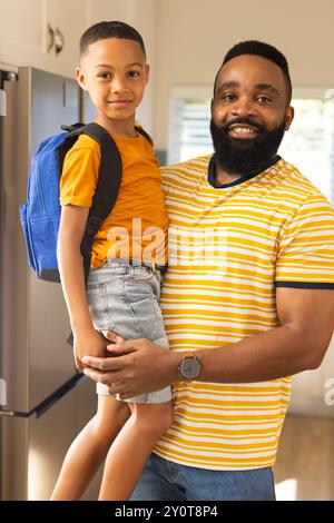 Padre che tiene il figlio con lo zaino, sorridente e pronto per la scuola in cucina Foto Stock