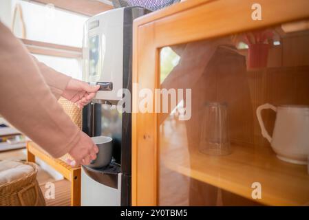 Una donna sta riempiendo una tazza da un distributore di refrigerazione dell'acqua. Il dispenser del refrigeratore d'acqua si trova su un ripiano in legno accanto a un armadio in vetro Foto Stock