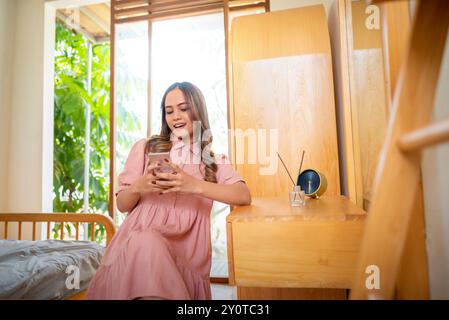 Una donna indonesiana con un vestito rosa è seduta su una sedia e guarda il suo telefono. La camera ha un letto e una scrivania in legno con un orologio e un vaso. Cen Foto Stock