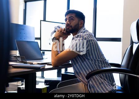 Pensando profondamente, un uomo indiano seduto alla scrivania con un laptop e un braccio robotico in ufficio Foto Stock