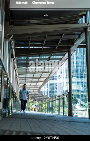 Hong Kong, Cina - 3 luglio 2024: Un uomo cammina su una passerella pedonale con una ringhiera di vetro. Un alto edificio con molte finestre e' sullo sfondo. Foto Stock