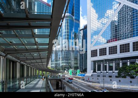 Hong Kong, Cina - 3 luglio 2024: Una passerella pedonale con ringhiere di vetro si affaccia su una trafficata via di Hong Kong. Grattacieli moderni fiancheggiano la strada, refle Foto Stock