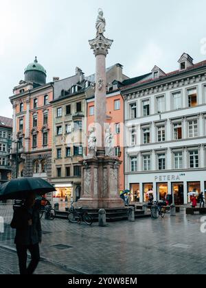 Una figura solitaria cammina sotto un ombrello in una vivace piazza della città, con una colonna decorativa e sculture circondate da affascinanti architetture e negozi. Foto Stock