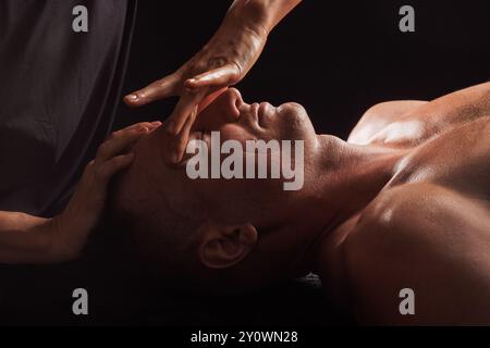 Primo piano di un uomo che riceve un massaggio facciale su uno sfondo scuro, le mani del massaggiatore premono delicatamente la fronte e. Foto Stock