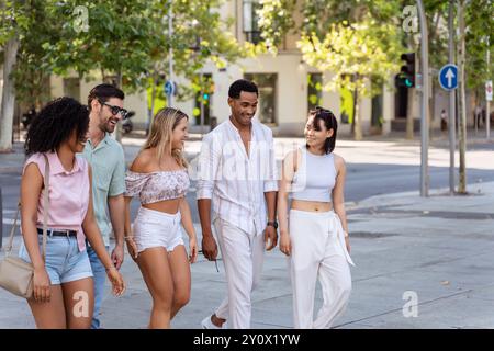Un gruppo variegato di cinque amici che si godono una passeggiata informale, ridendo e parlando in una giornata di sole in una strada alberata della città. Foto Stock