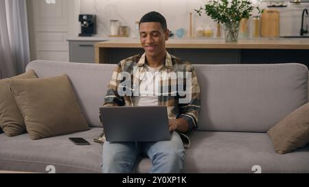 Felice sorridente afro-americano uomo etnico che lavora a casa lontano lavoro online studiando al computer portatile del divano che usa il computer sul divano maschio che usa il pc Foto Stock