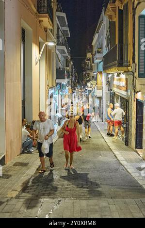 Sitges, Barcellona - 4 settembre 2024: Le persone camminano lungo una strada illuminata da luci a corda, circondata da edifici con balconi. Foto Stock