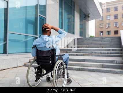 Uomo nero irritato in sedia a rotelle che non ha la possibilità di entrare nell'edificio senza rampa, all'aperto Foto Stock