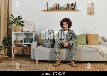 Una giovane donna afroamericana in equipaggiamento militare siede con cura nel suo soggiorno, pronta per essere schierata. Foto Stock