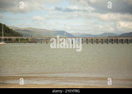 Piccola barca a vela di fronte al ponte ferroviario di Barmouth che attraversa l'estuario del Mawddach tra Fairbourne e Barmouth, Gwynedd, Galles del Nord, Regno Unito. Foto Stock