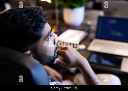 Pensando profondamente, l'uomo indiano seduto alla scrivania con un notebook e un tablet in ufficio Foto Stock