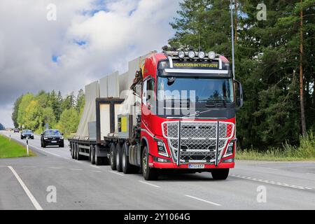 Veicoli e rimorchi Volvo FH rossi che trasportano componenti in calcestruzzo prefabbricato alti nel traffico stradale. Jokioinen, Finlandia. 8 agosto 2024. Foto Stock