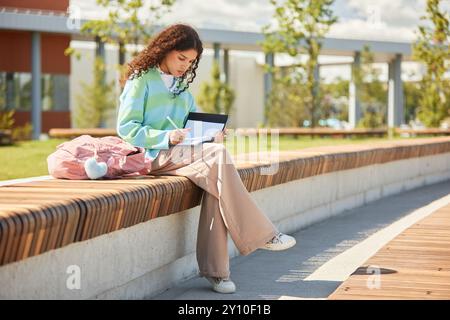 Studentessa riccia seduta sulla panchina con zaino posto accanto a una ragazza adolescente mentre scrive su un notebook che fa i compiti nel giorno di sole nel parco, scena di apprendimento informale all'aperto Foto Stock