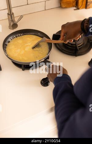 Cuocere uova strapazzate su fornello, uomo afroamericano che mescola con spatola di legno in cucina Foto Stock