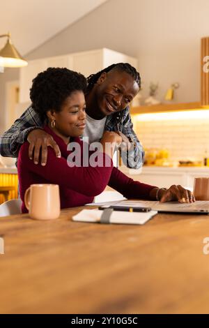 Uomo sorridente che abbraccia una donna afroamericana mentre lavora al laptop a casa Foto Stock