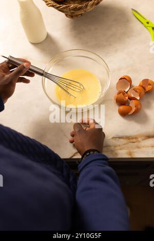 Montare le uova in un recipiente di vetro, un uomo afroamericano che prepara gli ingredienti per cucinare in cucina Foto Stock